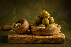 Picture of olive oil being drizzled over green olives within a wooden spoon, wooden bowl, and wooden cutting board